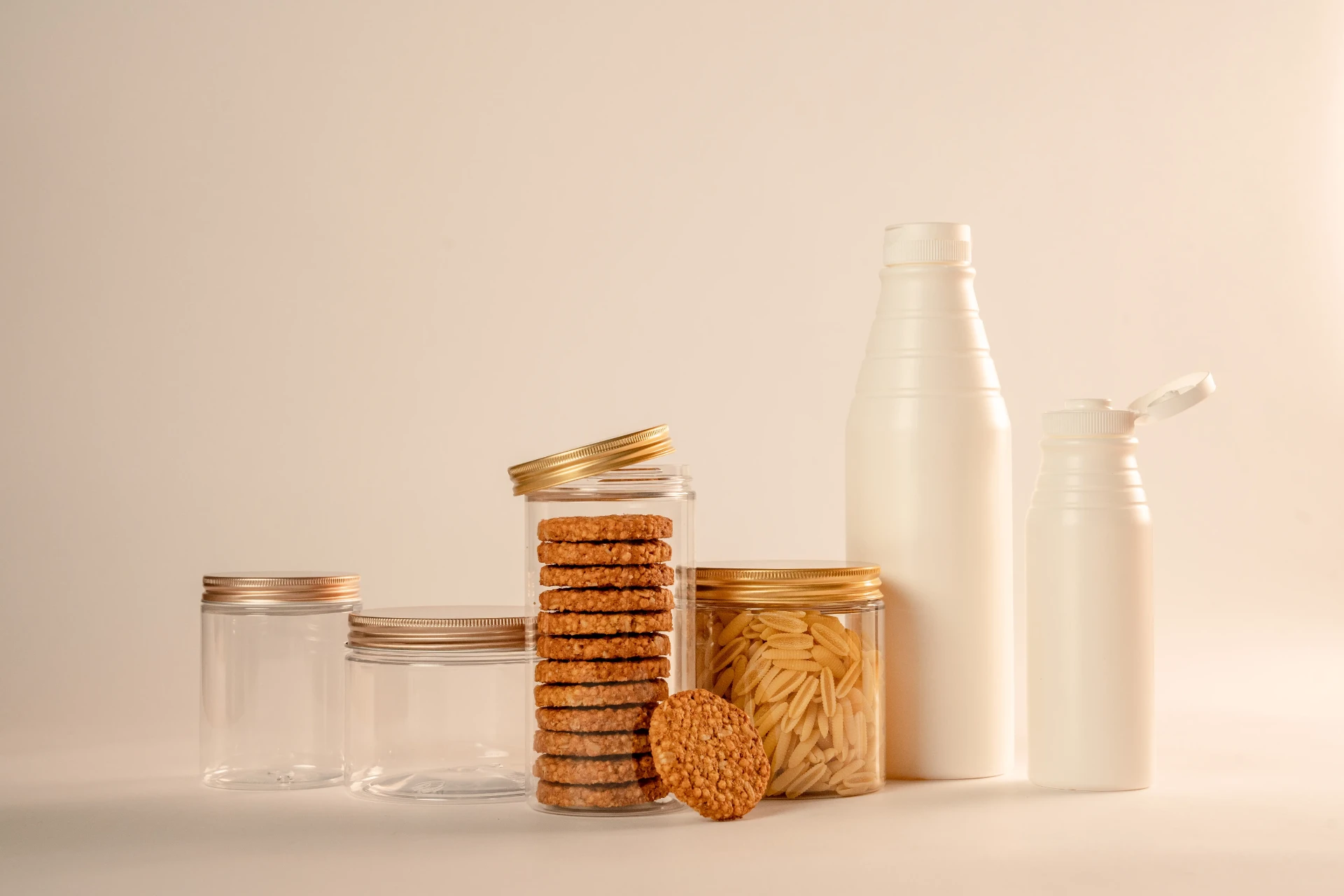 Barattoli trasparenti con capsula dorata contenenti biscotti e pasta, accanto a due flaconi bianchi per alimenti