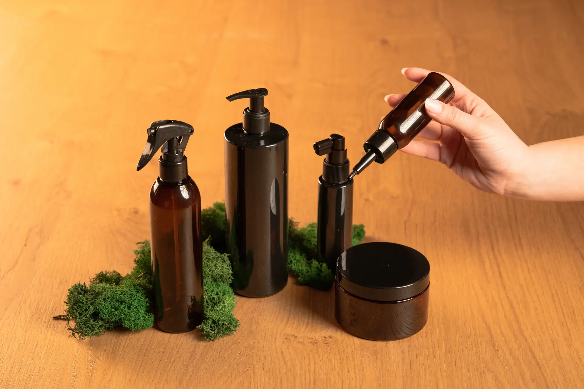 Una mano solleva un flacone ambrato con pompa da una linea di contenitori neri per capelli, su piano di legno con muschio