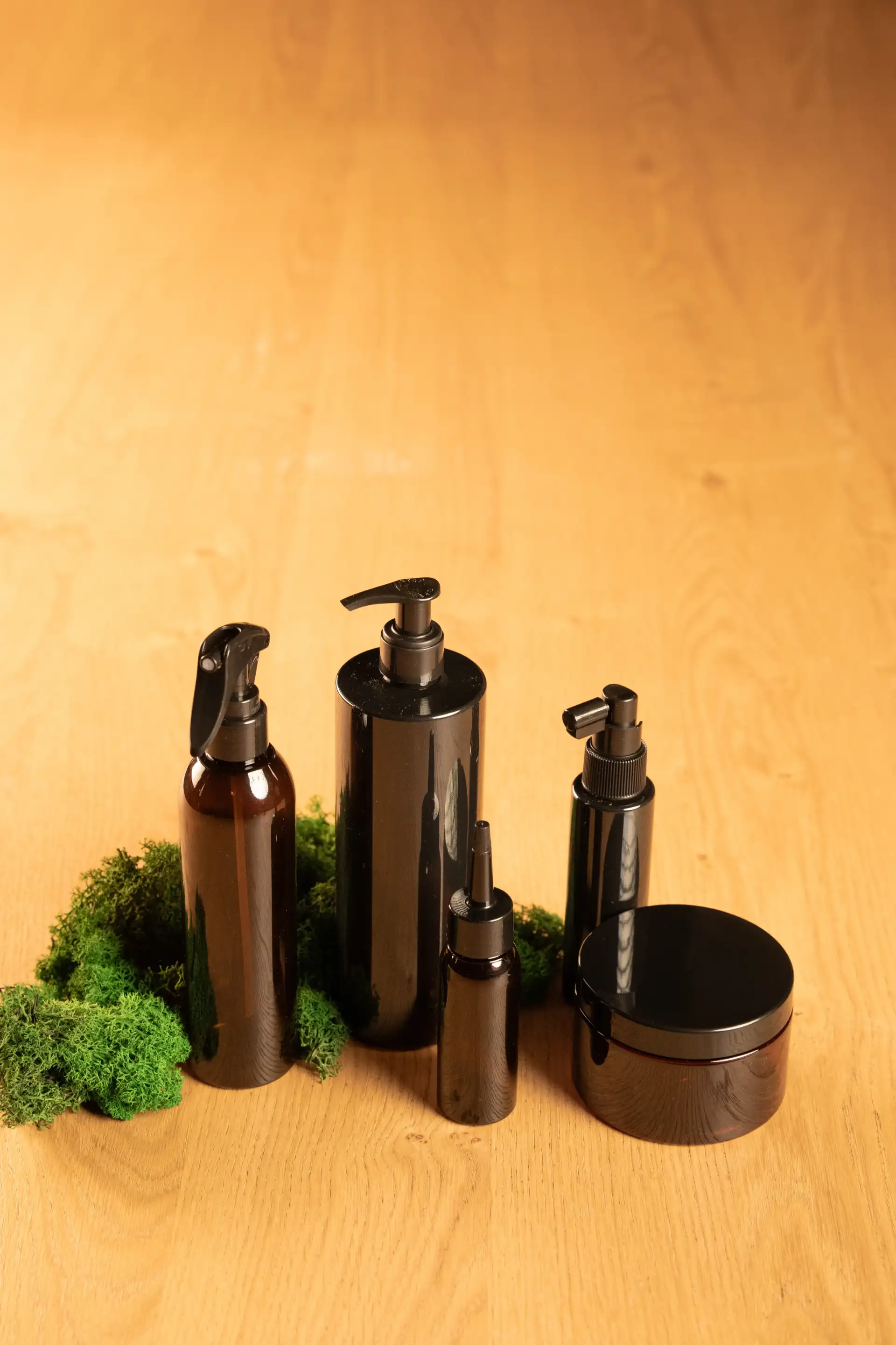 Cinque contenitori neri per la cura dei capelli — spray, dispenser a pompa, contagocce e vasetto — su un piano di legno chiaro con muschio