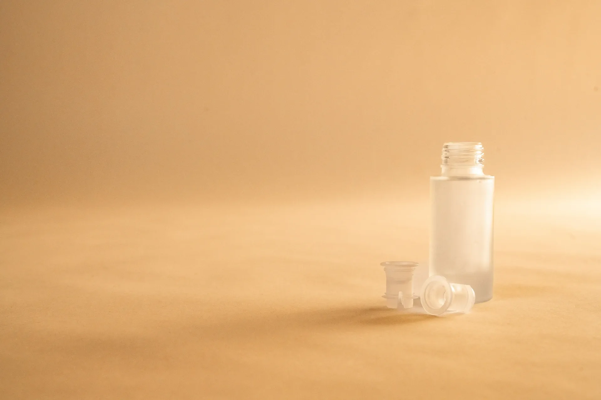 Flacone in vetro satinato aperto, con accanto due inserti in plastica traslucida, su fondo ocra
