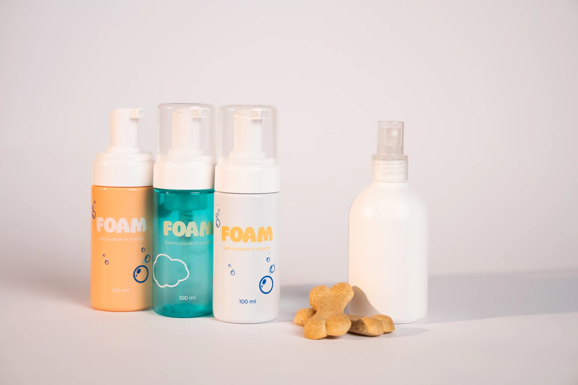 Tre erogatori di schiuma da 100 ml in arancione, verde acqua e bianco accanto a un flacone spray bianco e a biscotti per cani a forma di osso
