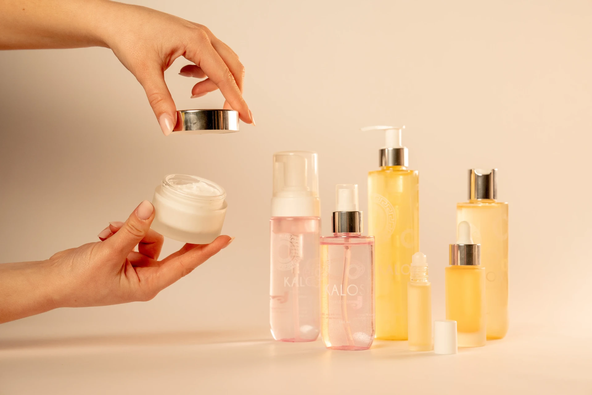 Mani che aprono un vasetto di crema in vetro satinato davanti a una linea di flaconi cosmetici rosa e ambra con pompe, spray e contagocce